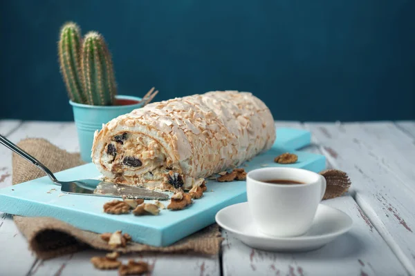 A cup of coffee meringue roll on a white vintage wooden kitchen table with burlap napkin. Meringue pie decorated with prunes and walnuts on blue cutting board.