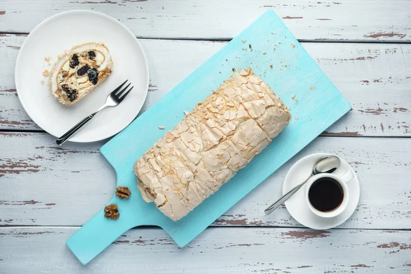A cup of coffee and meringue roll on a white vintage wooden kitchen table. Meringue pie decorated with prunes and walnuts on blue cutting board. Top view.