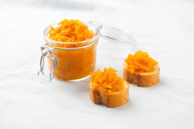 Beyaz masa örtüsü üzerinde iki parça Fransız ekmeği ve beyaz kumaş arka planında ev yapımı reçel olan bir kavanoz turuncu turuncu turşu.