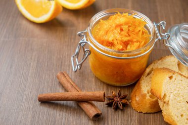 Tahta arka planda Fransız ekmeği, tarçın ve yıldız anason parçalarıyla dolu bir kavanoz turuncu konfeksiyon, ahşap masada birkaç parça ekmek bulunan ev yapımı portakal reçeli.