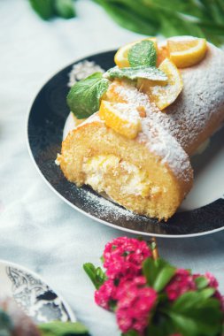 Limon meyvesi dilimleri ve nane yapraklarıyla süslenmiş bir dilim limonlu kek ve beyaz masa örtüsünde kırmızı çiçekler.