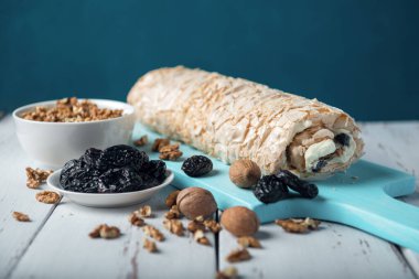 Beyaz ahşap mutfak masasında mavi bir kesme tahtasının üzerinde kuru erikli fındıklı krema rulosu. Kuru erik ve cevizli kremalı pasta ve kase..