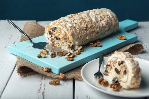 A piece of meringue roll with cake server and meringue roll on a white vintage wooden kitchen table with burlap napkin. Meringue pie decorated with prunes and walnuts on blue cutting board.