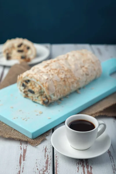 A cup of coffee meringue roll on a white vintage wooden kitchen table with burlap napkin. Meringue pie decorated with prunes and walnuts on blue cutting board.