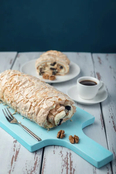 A cup of coffee meringue roll on a white vintage wooden kitchen table. Meringue pie decorated with prunes and walnuts on blue cutting board.