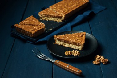 Siyah tabakta bir parça tart ve mavi ahşap masada doğranmış cevizli karamelli turta.