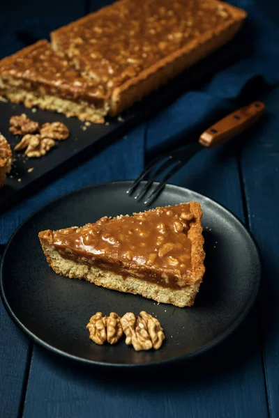 A piece of tart on the black plate and chopped walnut caramel pie on the blue wooden table