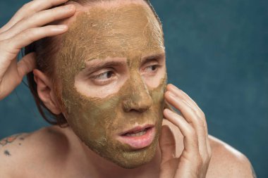 Yüzünde yeşil kilden maske olan ıslak uzun saçlı bir adamın spa prosedürünü beklerken yakın plan portresi..