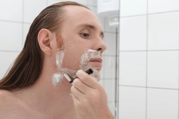 Serious tattooed man with long hair shaving his face in front of the bathroom mirror.