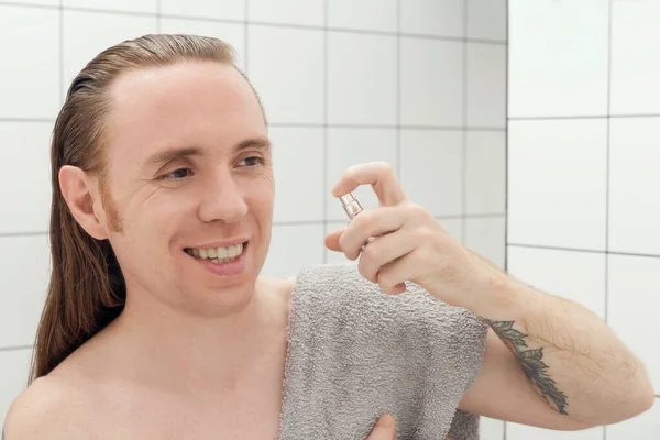 A man with tattoos and long hair sprays after-shave lotion in the bathroom