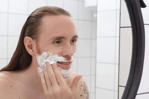 Brutal tattooed man with long hair with shaving foam on his face smiling in front of the bathroom mirror.