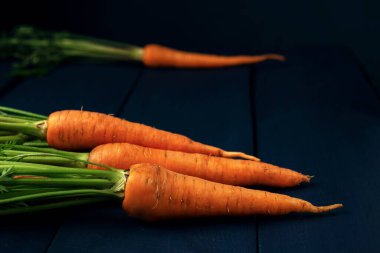 Mavi ahşap bir masada yeşil yapraklı bir avuç çiğ havuç. Koyu arkaplanda taze sebzeler. 