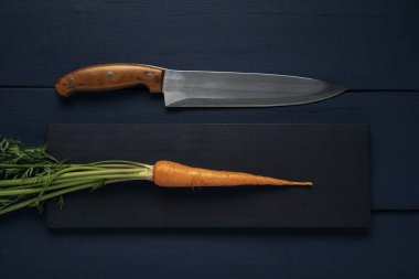 Tek bir çiğ havuç, yeşil yapraklar ve ahşap bir masada mutfak bıçağı. Üst görünüm.