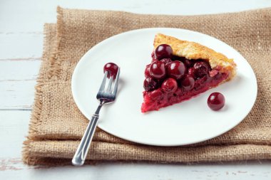 Olgun kırmızı kiraz dolgulu bir galet ve beyaz tabakta tatlı çatalı ve çuval bezi. Provence tarzı ahşap arka planda ev yapımı tatlı turta. 
