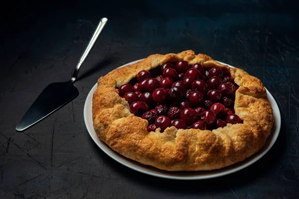 Galette with ripe red cherry filling and cake server on dark blue background. Homemade sweet open pie. Bakery product.