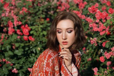 Kırmızı güllerin yanında poz veren güzel, hayalperest bir kadın. Sert güneş ışığı olan romantik bir kadının açık hava portresi..