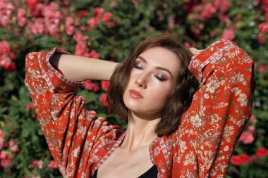 Gül bahçesinde poz veren kapalı gözlü güzel bir kadın. Sert güneş ışığıyla romantik bir kızın açık hava moda portresi..