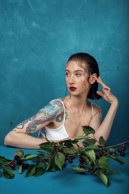 Genç manken mavi arka planda kiraz dallı masada oturuyor.