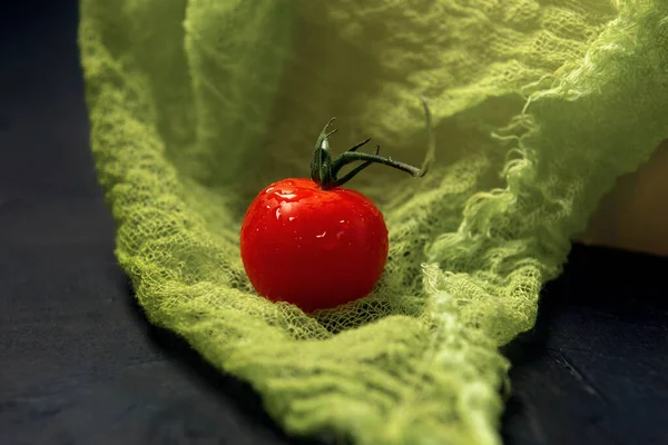 A single red wet cherry tomato on a green gauze background.