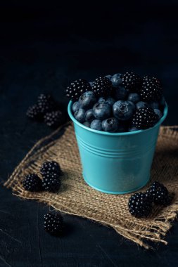 Çuval bezinde yabanmersini ve böğürtlenli mavi kova ve koyu dokulu arka plan..