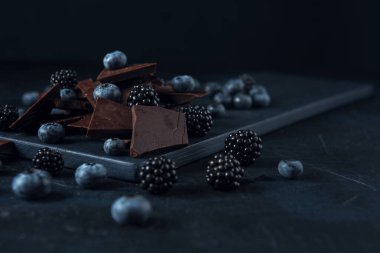 Böğürtlen ve böğürtlen, siyah kesim tahtasında çikolata parçaları ve koyu dokulu arka plan.. 