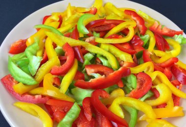 Dilimlenmiş dolmalık biber, kırmızı, sarı ve yeşil, siyah arka planda beyaz bir tabakta, yukarıdan yakın çekim görüntüsü