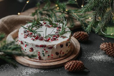 Beyaz peynir kremalı Noel pastası, yaban mersini ve biberiyeyle süslenmiş. Köknar dalları ve çelenklerin arka planında yılbaşı hediyesi.