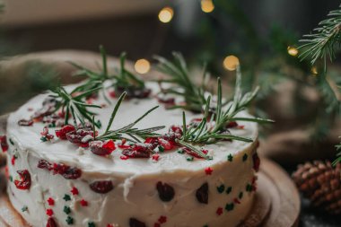 Beyaz peynir kremalı Noel pastası, yaban mersini ve biberiyeyle süslenmiş. Köknar dalları ve çelenklerin arka planında yılbaşı hediyesi.