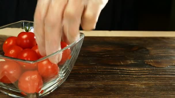Chef com as mãos leva tomates cereja de tigela de vidro transparente e corta vegetais em metades com faca de cozinha na placa de corte escuro. Close-up . — Vídeo de Stock