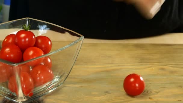Koki dengan tangannya mengambil tomat ceri dari mangkuk kaca transparan dan memotong sayuran menjadi dua bagian dengan pisau dapur di papan potong cahaya. Close-up. — Stok Video