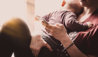 Kadın milenyum kuşağı bir bebek ve akıllı telefon tutuyor. Genç bir kadın küçük bir çocuğa bakıyor ve akıllı telefon kullanıyor. Telefon sosyal medyada ya da internet üzerinden iş için kullanılır.