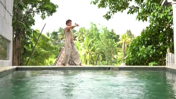 衣装のハンサムな男はプールの近くの夏のテラスで屋外で気功演習を練習します 癒しと精神的健康 バリ島 インドネシア — ストック動画
