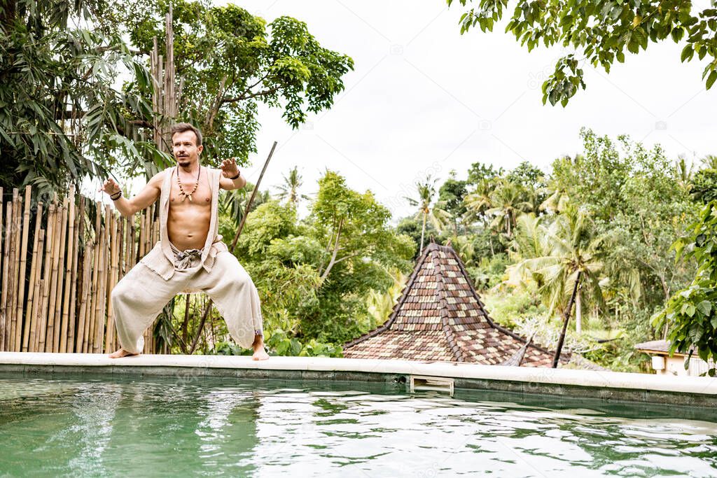 Handsome man in costume practice qigong exercises outdoors in summer terrace near the pool. Healing and spiritual Wellness, Bali, Indonesia