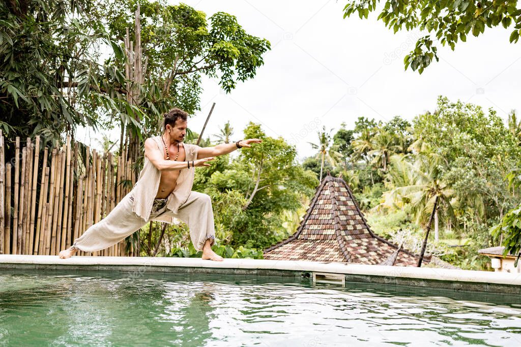 Handsome man in costume practice qigong exercises outdoors in summer terrace near the pool. Healing and spiritual Wellness, Bali, Indonesia