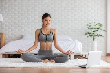 Laptop talimi yapan çekici Asyalı kadın yoga nilüferleri evde meditasyon ve rahatlama, evde sağlık ve egzersiz, COVID-19 salgını nedeniyle kendini izole etme ve kondisyon kursuna poz veriyor.