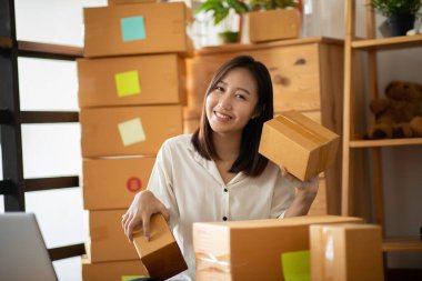 Evde online alışveriş için dizüstü bilgisayarla çalışan genç Asyalı kadınların KOBİ girişimcisi, evde paketleme için kutusuyla Neşeli ve Mutlu, Business Online ve Online alışveriş için kendi İş Kurma 