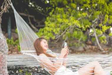 Beyaz bikini giyen, cep telefonu kullanan, Tayland 'da tropikal ada sahillerinde hamakta uzanan mutlu Asyalı kadınların yaz tatili Tayland' da seyahat, yaz tatilleri.