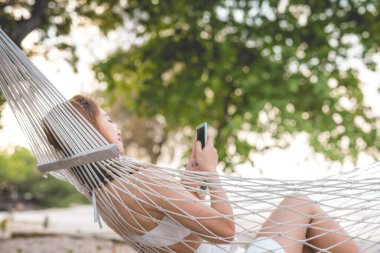 Beyaz bikini giyen, cep telefonu kullanan, Tayland 'da tropikal ada sahillerinde hamakta uzanan Tayland' da seyahat eden mutlu Asyalı kadınların yaz tatili.