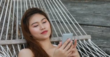 Beyaz bikini giyen, cep telefonu kullanan, Tayland 'da tropikal ada sahillerinde hamakta uzanan mutlu Asyalı kadınların yaz tatili Tayland' da seyahat, yaz tatilleri.