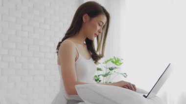 Güzel, çekici, Asyalı genç bir kadın yatakta oturuyor ve bilgisayarını sosyal medya ve online alışveriş için kullanıyor tatilde rahat bir yatak odasında dinleniyor, yatak odasında yaşam tarzı kavramını kullanıyor.
