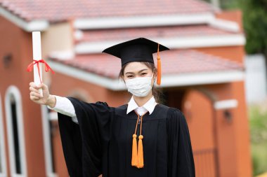 Mezun olmuş bir kadın, virüsü korumak için koruyucu elbise ve tıbbi maske takıyor. Virüs salgını ve COVID-19 virüsü salgını nedeniyle diploma ya da sertifikalı. Mezuniyet gününün yeni normal vakası.