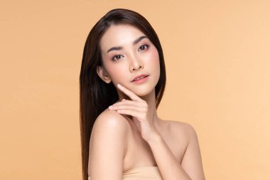 Güzel Asyalı genç bir kadın yumuşak yanaklı gülüşüne dokunuyor taze tenli mutlu ve neşeli, olumlu duygusal, bej arka planda soyutlanmış, güzellik estetiği ve yüz bakımı kavramında.