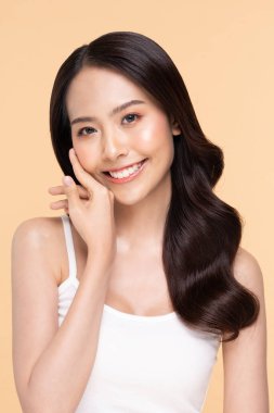 Güzel Asyalı genç bir kadın yumuşak yanaklı gülüşüne dokunuyor taze tenli mutlu ve neşeli, olumlu duygusal, bej arka planda soyutlanmış, güzellik estetiği ve yüz bakımı kavramında.