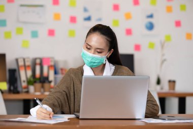 Yüz maskesi takan mutlu Asyalı iş kadını modern ofiste ya da Ortak Çalışma Alanı, İş Kuruluşu Konsepti 'nde çalışıyor.