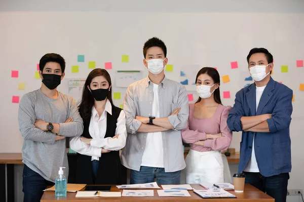 Group of Asian business men leader standing wearing mask smile cross arm confidence with Business team at Co-Working space Due Covid-19 flu pandemic and protection,Small Business Startup Concept