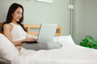Güzel, çekici, Asyalı genç bir kadın yatağa uzanıyor ve bilgisayarını sosyal medya ve online alışveriş için kullanıyor tatillerde rahat yatak odasında dinleniyor, yatak odasında yaşam tarzı kavramını kullanıyor. 