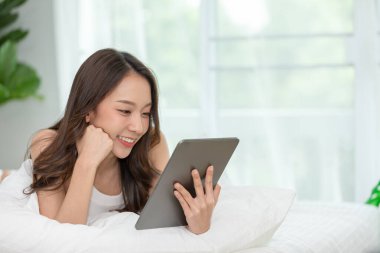 Güzel Asyalı kadın gülümseyip evdeki yatağında bilgisayar tableti kullanıyor, sosyal medya ve multimedya kullanarak tatilde rahatlıyor.