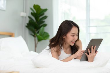 Güzel Asyalı kadın gülümseyip evdeki yatağında bilgisayar tableti kullanıyor, sosyal medya ve multimedya kullanarak tatilde rahatlıyor.