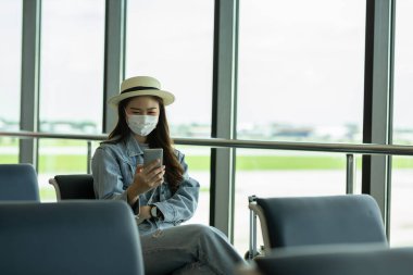 Covid-19 gribi salgını ve korunması nedeniyle havaalanında cep telefonu kullanan Asyalı genç bir kadın yolcu.