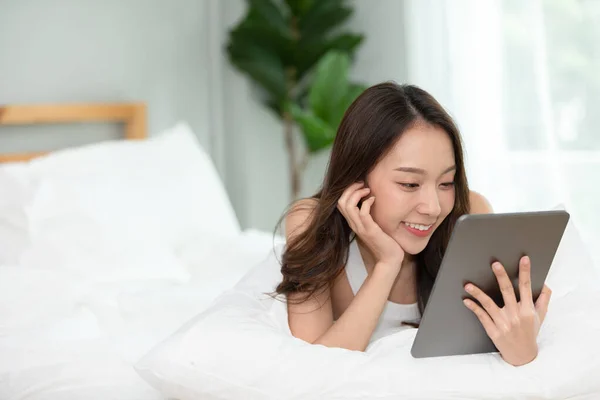 Beauty Asian woman smile and using computer tablet on her bed at home,using social media and multimedia on tablet relax in holiday